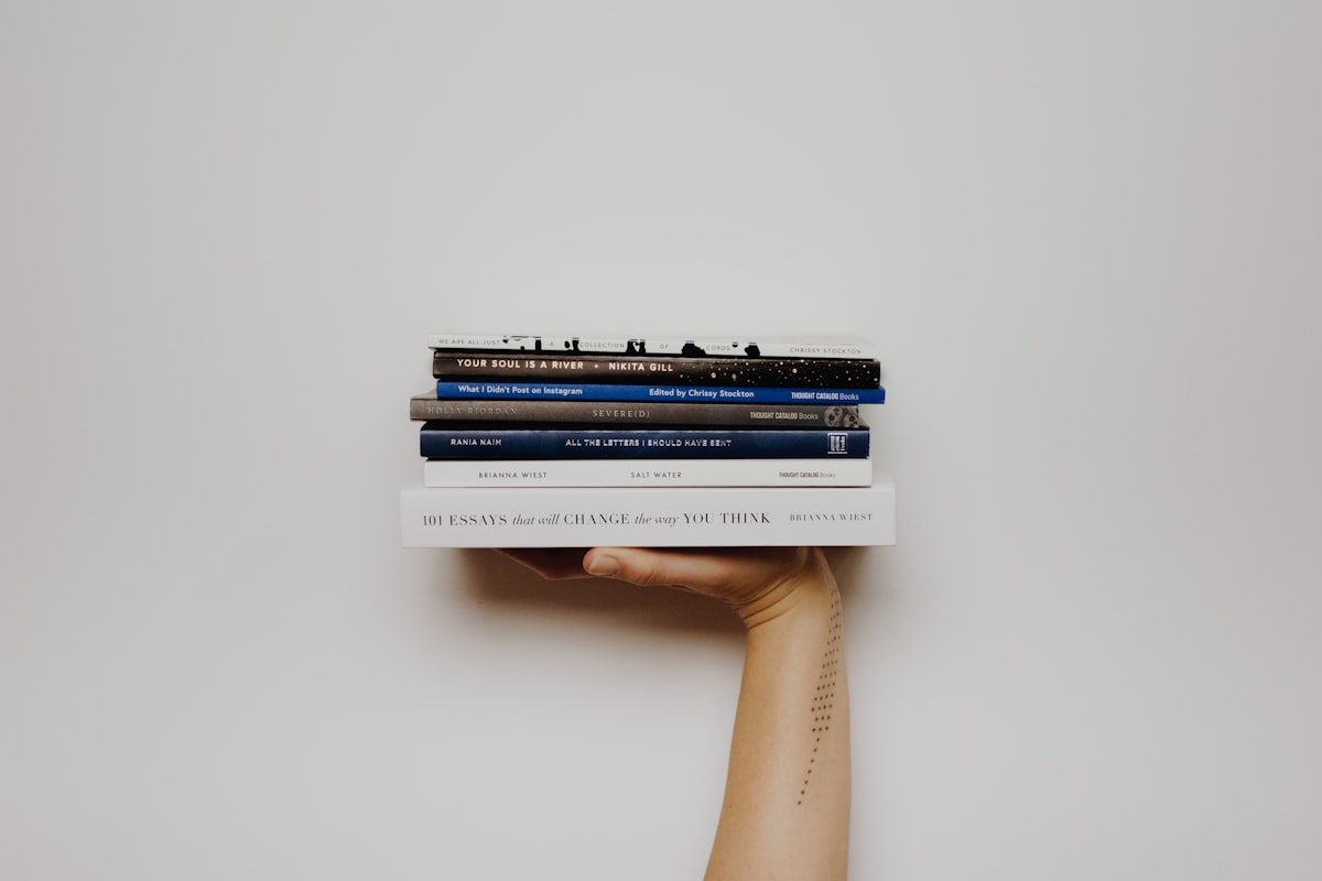 Hand and forearm holding a stack of seven paperback books against a plain off-white wall